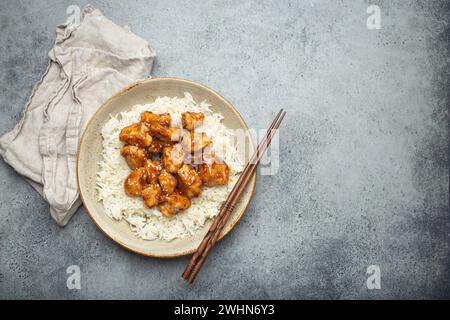 Chinesisches süß-saures, klebriges Hühnchen mit Sesamsamen und Reis auf Keramikplatte mit Essstäbchen von oben auf grauem rustikalem Stein Stockfoto