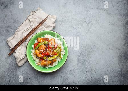 Asiatisches süß-saures Hähnchen mit Gemüse, gebraten und Reis in Keramikschale mit Essstäbchen von oben auf graue rustikale Str. Stockfoto