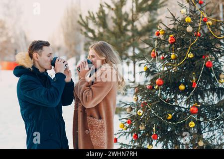 Verliebte Paare trinken Kaffee neben dem geschmückten Weihnachtsbaum im Winterwald Stockfoto