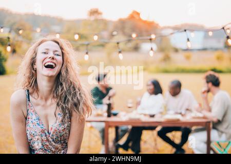 Lachende schöne junge Frau mit lockigen blonden Haaren in einem Sommerkleid mit einer Gruppe von Freunden im Hintergrund, die ein Outdo haben Stockfoto