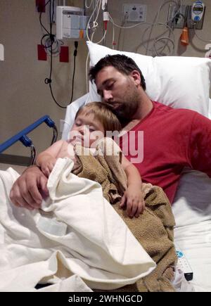 Ein müder Vater kuschelt sich mit seinem Kleinkind-Sohn in einem Krankenhausbett, nachdem das Kind operiert wurde Stockfoto