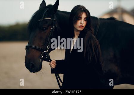 Junge Frau in Schwarz mit ihrem Pferd bei Sonnenuntergang draußen. Stockfoto