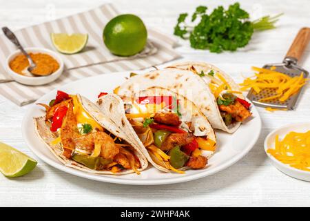 Nahaufnahme von gebackenen Tex-mex-Hühnchen-Fajitas mit gemischtem süßem Pfeffer, Zwiebeln, Sauerrahm, geriebenem Käse und weißen Maistortillas auf weißer Platte auf Holz Stockfoto