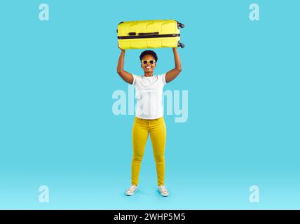 Porträt einer Frau in voller Länge mit gelbem Koffer über dem Kopf, lächelnd auf türkisfarbenem Hintergrund. Stockfoto