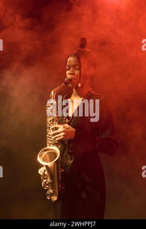 Vertikales Porträt einer schwarzen jungen Frau, die Saxophon auf der Bühne in einem Nachtclub mit Raucheffekten spielt Stockfoto