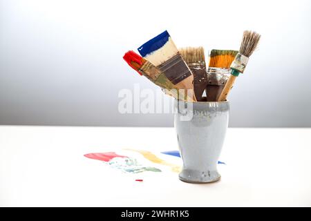 Mehrere benutzte Pinsel mit verschiedenen Farben in einem Keramikbecher und Farbflecken auf dem weißen Tisch vor grauem Hintergrund, Stockfoto
