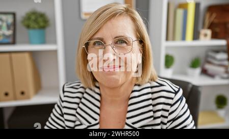 Eine lächelnde Frau mittleren Alters mit Brille in einem gestreiften Blazer sitzt in einem modernen Büro Stockfoto