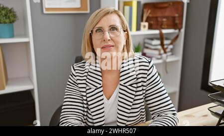 Selbstbewusste reife Frau mit Brille in einem gestreiften Blazer am Schreibtisch Stockfoto