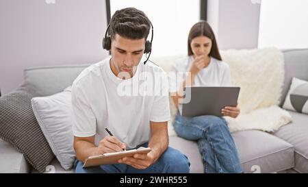 Ein Mann mit Kopfhörern, der sich auf das Schreiben konzentriert, während eine Frau in einem modernen Wohnzimmer an einem Laptop arbeitet. Stockfoto