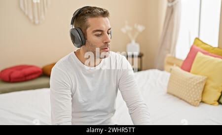 Ein nachdenklicher junger Mann mit Kopfhörern sitzt zu Hause in einem weißen Hemd auf dem Bett, beschäftigt sich mit Freizeit oder Arbeit in einer ruhigen Schlafzimmerumgebung. Stockfoto