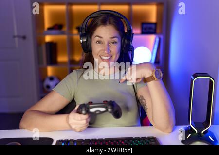 Schöne brünette Frau, die Videospiele mit Kopfhörern spielt und selbstbewusst mit einem Lächeln auf dem Gesicht aussieht, sich mit den Fingern stolz und glücklich zeigt. Stockfoto