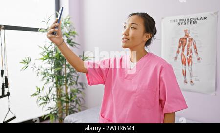 Eine junge asiatische Frau in einem rosafarbenen Gestrüpp macht ein Selfie in einer Klinik mit einem Poster für das Muskelsystem im Hintergrund. Stockfoto