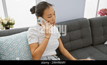Eine junge asiatische Frau, die in einem modernen Wohnzimmer bequem auf einem Sofa sitzt und ein Telefongespräch führt. Stockfoto