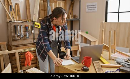 Eine fokussierte junge Frau skizziert Entwürfe in ihrer Tischlerei, umgeben von Werkzeugen und Holz. Stockfoto