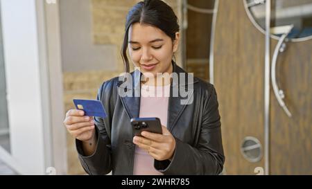Eine junge hispanische Frau prüft ihre Kreditkarte und ihr Smartphone auf einer Straße der Stadt und verkörpert urbane Eleganz und Konnektivität. Stockfoto
