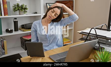 Überarbeitete, aber robuste, junge, schöne hispanische Frau dehnt den Hals und arbeitet unermüdlich auf dem Touchpad im Herzen des Büros Stockfoto