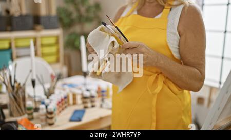 Wunderschöne blonde Künstlerin mittleren Alters, die im gemütlichen Kunststudio während des Kurses mit Leidenschaft Pinsel reinigt Stockfoto