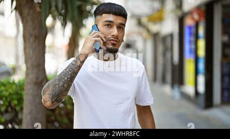 Cooler, tätowierter junger latino-Mann, der ernsthaft in Telefongespräche vertieft ist, draußen auf der sonnendurchfluteten Stadtstraße steht und entspannte Vibes ausstrahlt, während er s verwendet Stockfoto
