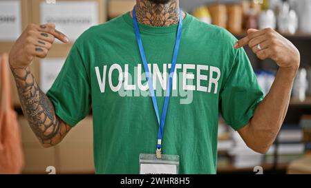 Hübscher junger lateinmann, tätowiert und arbeitet, selbstlos auf seine Freiwilligenuniform im belebten Lager des Wohltätigkeitszentrums gerichtet, stehend Stockfoto