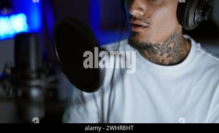 Hübscher junger lateinmann, ein tätowierter Musiker, vertieft in sein musikalisches Hobby, singt sein gefühlvolles Lied in einem schwach beleuchteten Musikstudio und gießt Sse Stockfoto