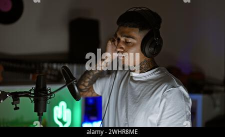 Hübscher junger lateinmann, ein tätowierter Musiker, vertieft in sein musikalisches Hobby, singt sein gefühlvolles Lied in einem schwach beleuchteten Musikstudio und gießt Sse Stockfoto