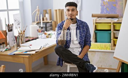 Tätowierter junger lateinamerikanischer Mann, ein vertiefter Künstler, der mit ernstem Gesicht in einem Kunststudio an seiner Staffelei sitzt, mit Pinsel in der Hand Stockfoto