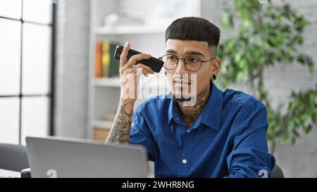Konzentrierter lateinamerikanischer Arbeiter, Porträt eines gutaussehenden, tätowierten Geschäftsexperten, ernst und vertieft, der in seinem Büro arbeitet und einer dringenden Sprachnachricht zuhört Stockfoto