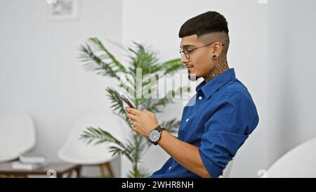 Hübscher junger lateinmann, tätowiert und ernst, vertieft in sein Smartphone, während er in einem Wartezimmerstuhl saß und die Zeit vor seinem Bu wegschickte Stockfoto