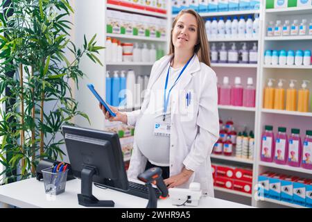 Junge schwangere Apothekerin lächelt selbstbewusst mit Computer und Touchpad in der Apotheke Stockfoto