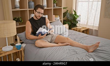 Junger hispanischer Mann mit Brille, der Buch liest, sitzt im Schlafzimmer auf dem Bett Stockfoto