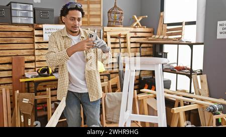 Ein junger erwachsener Mann mit lockigen Haaren macht ein Foto in einer Tischlerei, umgeben von Holz, Werkzeugen und Möbeln. Stockfoto