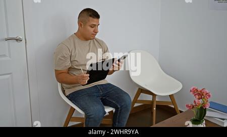 Ernsthafter junger lateinmann, der entspannt drinnen auf einem Stuhl sitzt, vertieft in das Schreiben wichtiger Notizen auf die Zwischenablage, während er gleichzeitig online über g schreibt Stockfoto