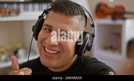 Ein hübscher junger hispanischer Kerl, ein professioneller Musiker, der in einem Musikstudio positiv mit der Geste nach oben drückt und lächelt, als er sich in seinem melodischen hobb verirrt Stockfoto