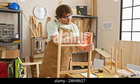 Ein fokussierter junger Mann mit Bart, Schutzbrille und Schürze, poliert einen Holzstuhl in einer gut ausgestatteten Tischlerei. Stockfoto