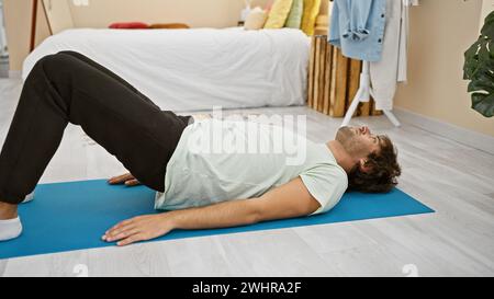Ein junger bärtiger Mann entspannt sich auf einer Yogamatte in einem hellen Schlafzimmer mit moderner Einrichtung und Zimmerpflanzen, die Wellness und Ruhe verkörpern. Stockfoto