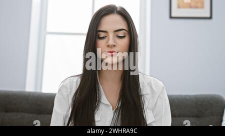 Eine ruhige junge hispanische Frau in einer weißen Bluse meditiert drinnen und bietet eine friedliche, attraktive Porträtszene. Stockfoto