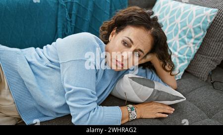 hispanische Frau mittleren Alters mit lockigem Haar, die kontemplativ auf einer Couch in einem gemütlichen Wohnzimmer zu Hause liegt und reife Schönheit ausstrahlt. Stockfoto