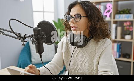 Reife hispanische Frau Podcasts aus einem hellen Radiostudio, die in ein Mikrofon spricht, während sie Kopfhörer trägt. Stockfoto