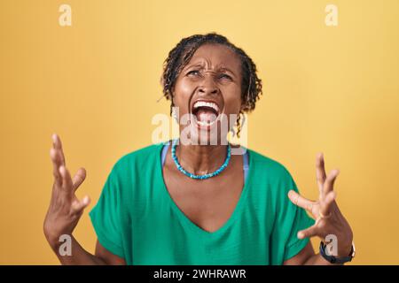Afrikanische Frau mit Dreadlocks, die über gelbem Hintergrund stehen, verrückt und verrückt schreien und schreien, mit aggressivem Ausdruck und erhobenen Armen. Frustrati Stockfoto