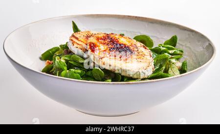 Gegrillter Ziegenkäse auf einem Bett aus frischen Spinatblättern, serviert in einer Keramikschale auf weißem Hintergrund. Stockfoto