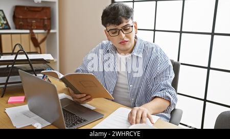 Ein junger Mann in Brille liest ein Notizbuch an einem überfüllten Schreibtisch mit einem Laptop und Dokumenten. Stockfoto