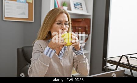 Reife kaukasische Frau genießt Kaffee in einem modernen Büro-Innenraum, die ein professionelles Ambiente am Arbeitsplatz darstellt. Stockfoto