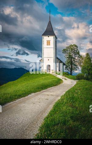 Frühlingslandschaft und traditionelle Kirche auf dem Hügel. Fantastischer Ort mit ländlicher Straße und St. Primoz Kirche im Hintergrund, Jamnik Dorf, Slowenien, EU Stockfoto