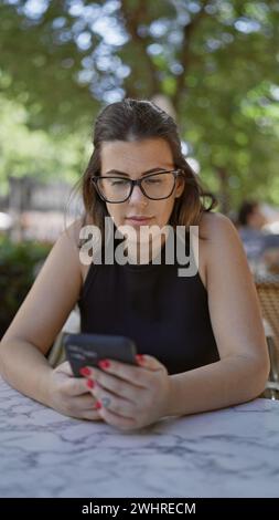 Lächelnde, schöne hispanische Frau, die fröhlich das Smartphone benutzt, während sie auf der Außenterrasse des Cafés sitzt, einen Kaffee genießt, mit dem digitalen verbunden ist Stockfoto