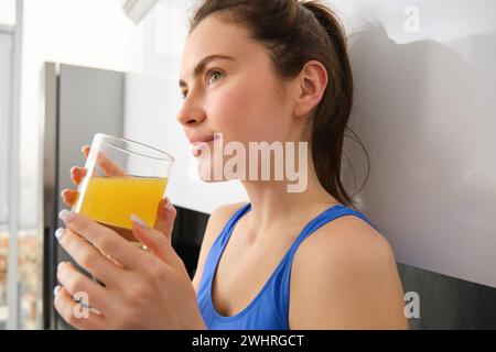 Nahaufnahme Porträt einer schönen jungen Frau, die wegschaut, Orangensaft trinkt, frisch im Glas, in der Küche steht, Verschleißauftrag Stockfoto