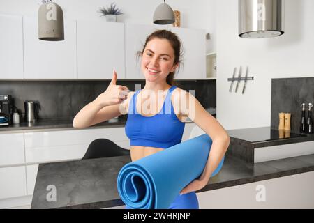 Schöne junge Frau, zeigt Daumen nach oben, hält Gummi-Yoga-Matte, macht Fitness-Übungen von zu Hause, Gym im Wohnzimmer, empfiehlt Stockfoto