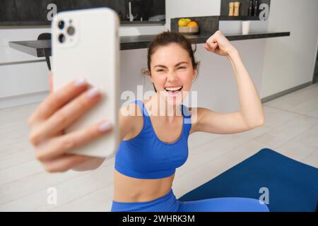 Das junge, energiegeladene Mädchen macht zu Hause Sport, zeigt ihre Muskeln am Arm und macht Selfie auf dem Smartphone, posiert auf der Fitness-Gummimatte Stockfoto