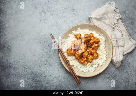 Chinesisches süß-saures, klebriges Hühnchen mit Sesamsamen und Reis auf Keramikplatte mit Essstäbchen von oben auf grauem rustikalem Stein Stockfoto