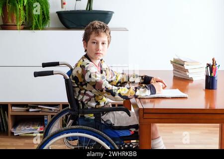 Ein kleiner Junge im Rollstuhl sitzt konzentriert an einem Schreibtisch, beschäftigt mit seinen Hausaufgaben. Stockfoto