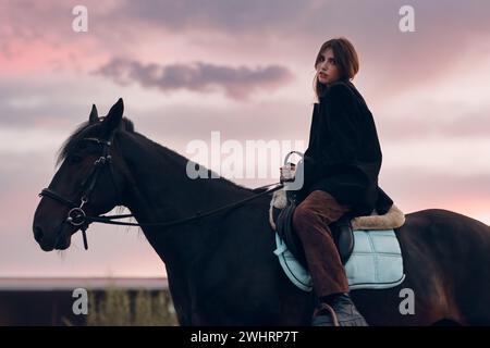 Junge Frau in Schwarz mit ihrem Pferd bei Sonnenuntergang draußen. Stockfoto